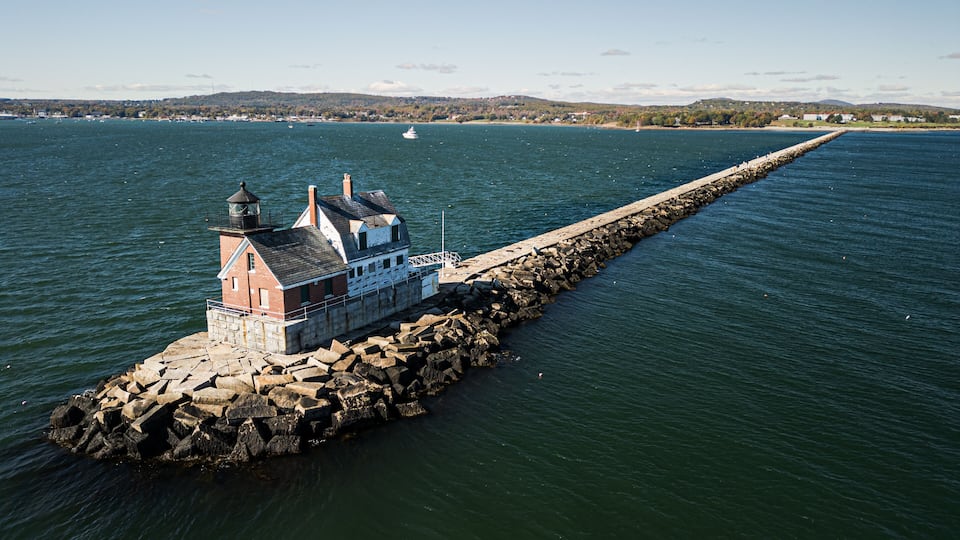 A lighthouse on a rocky pier extending into a calm sea, with a distant shoreline and hills.