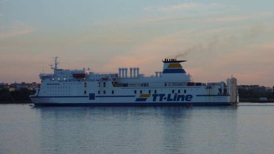 A large white and blue ferry with the name "TT-Line" on its side, sailing on calm waters during sunset.