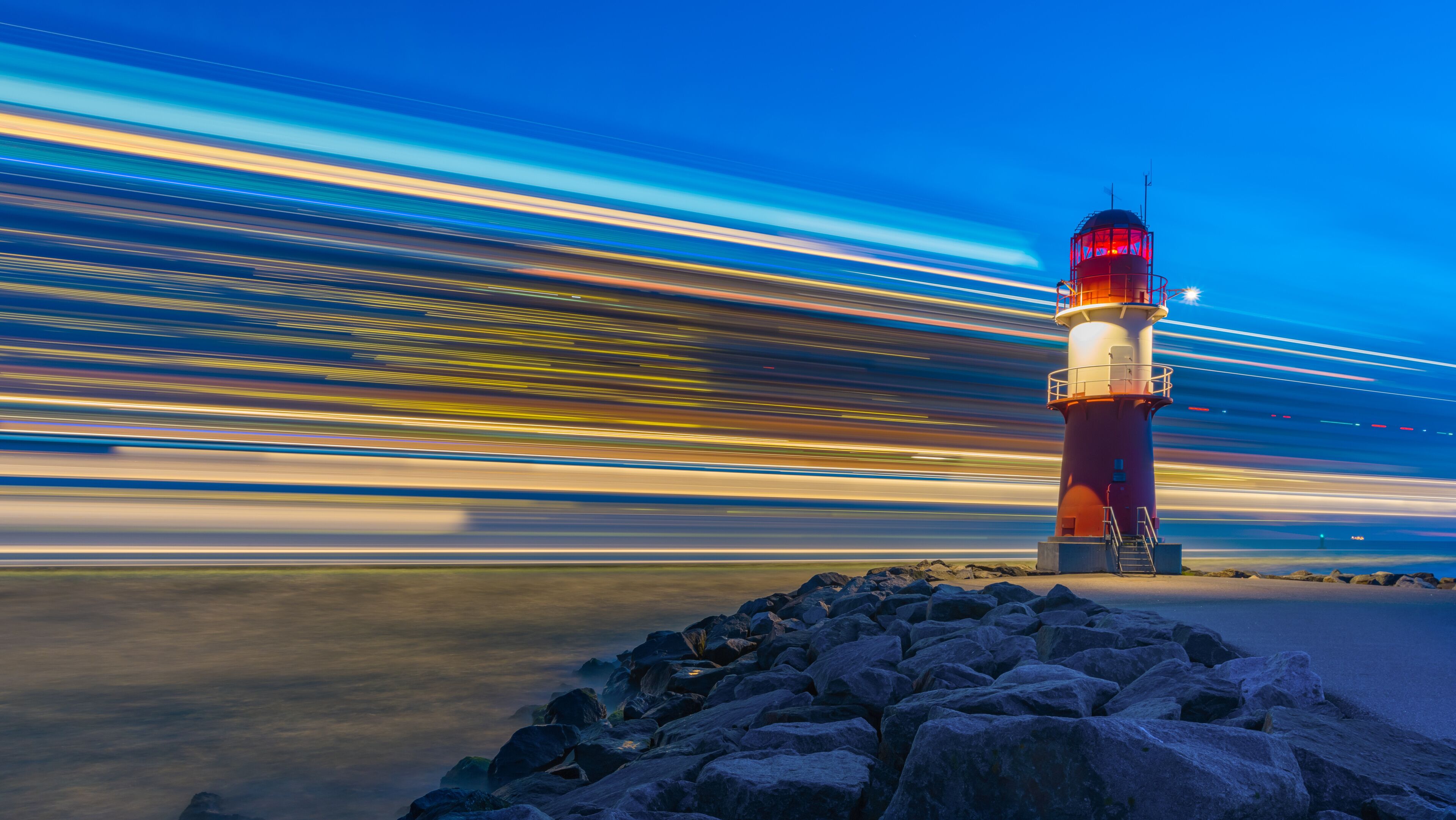 Un faro con la parte superior roja y la base blanca, rodeado de rocas y estelas de luz producto de una exposición prolongada.