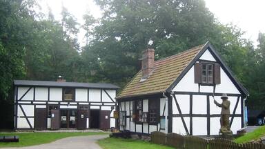 A traditional half-timbered house with a thatched roof, a statue of a figure, and a paved driveway.