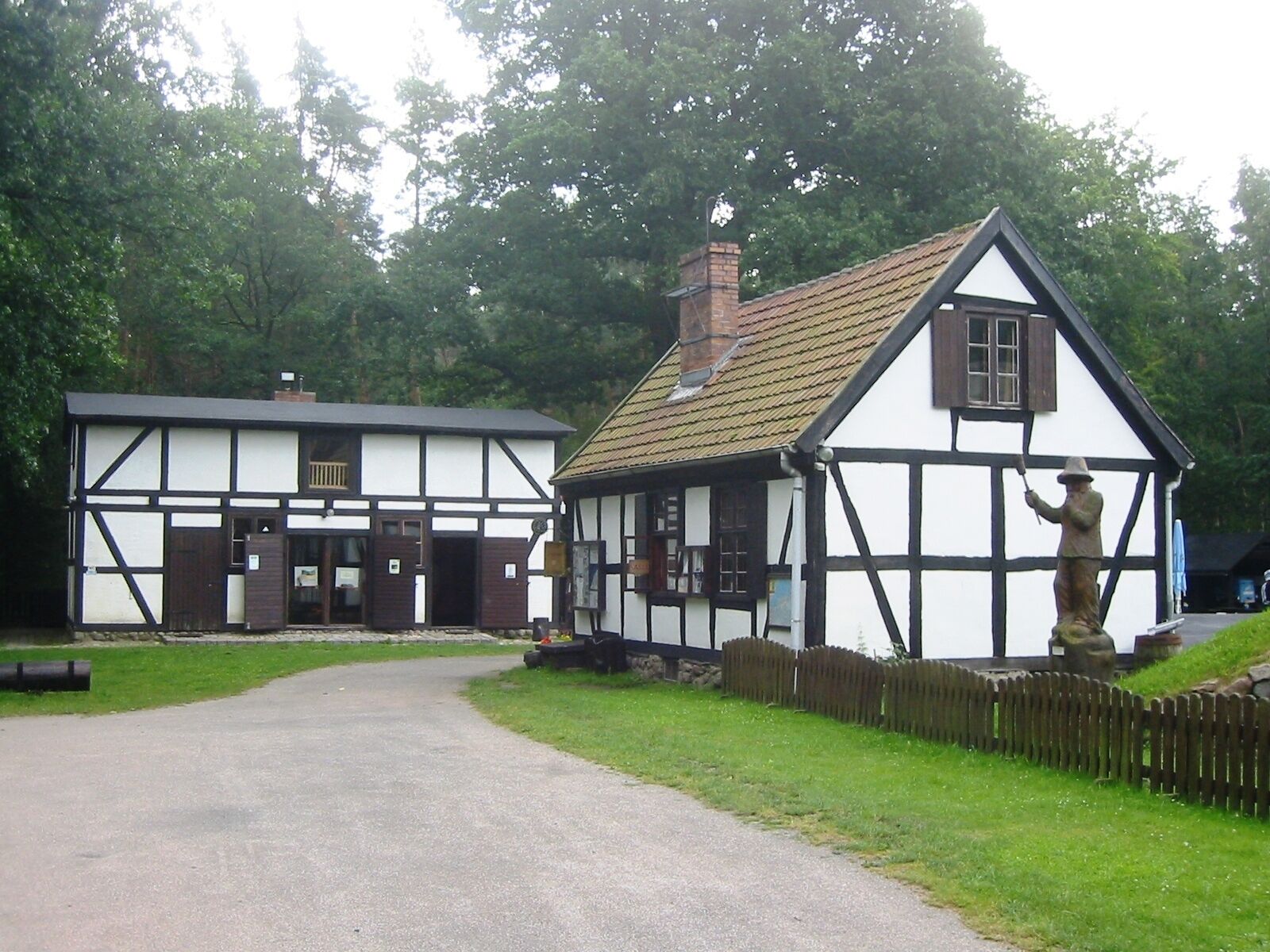 A traditional half-timbered house with a thatched roof, a statue of a figure, and a paved driveway.