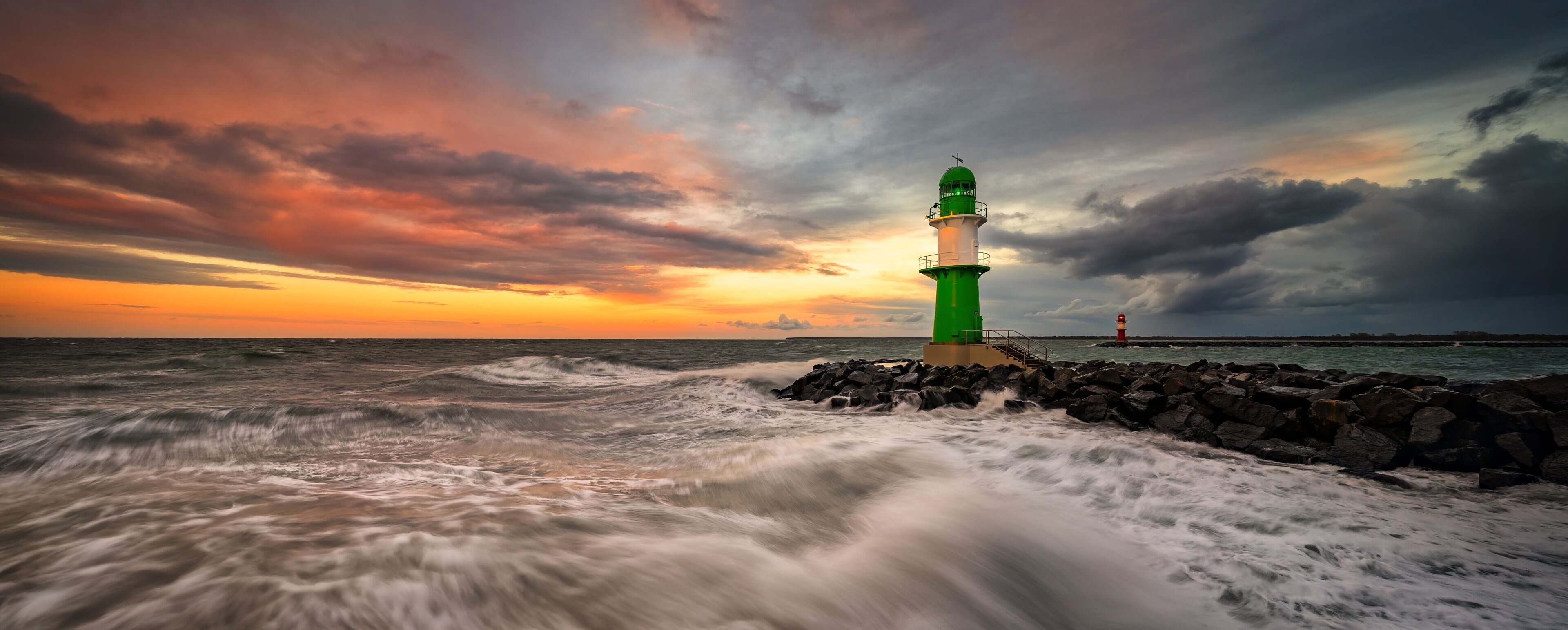 A green lighthouse by the sea during sunset with waves crashing against rocks.