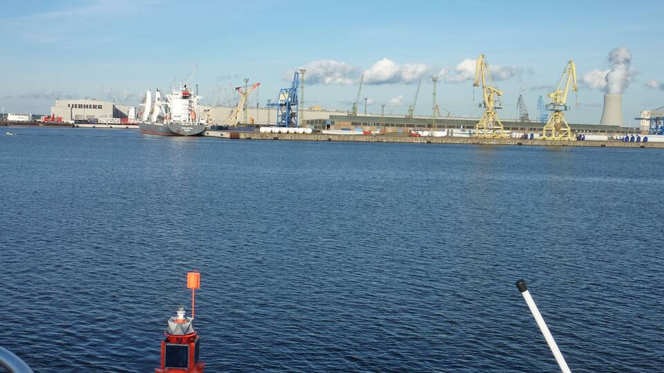 A large cargo ship docked at a port with industrial cranes and a refinery in the background.