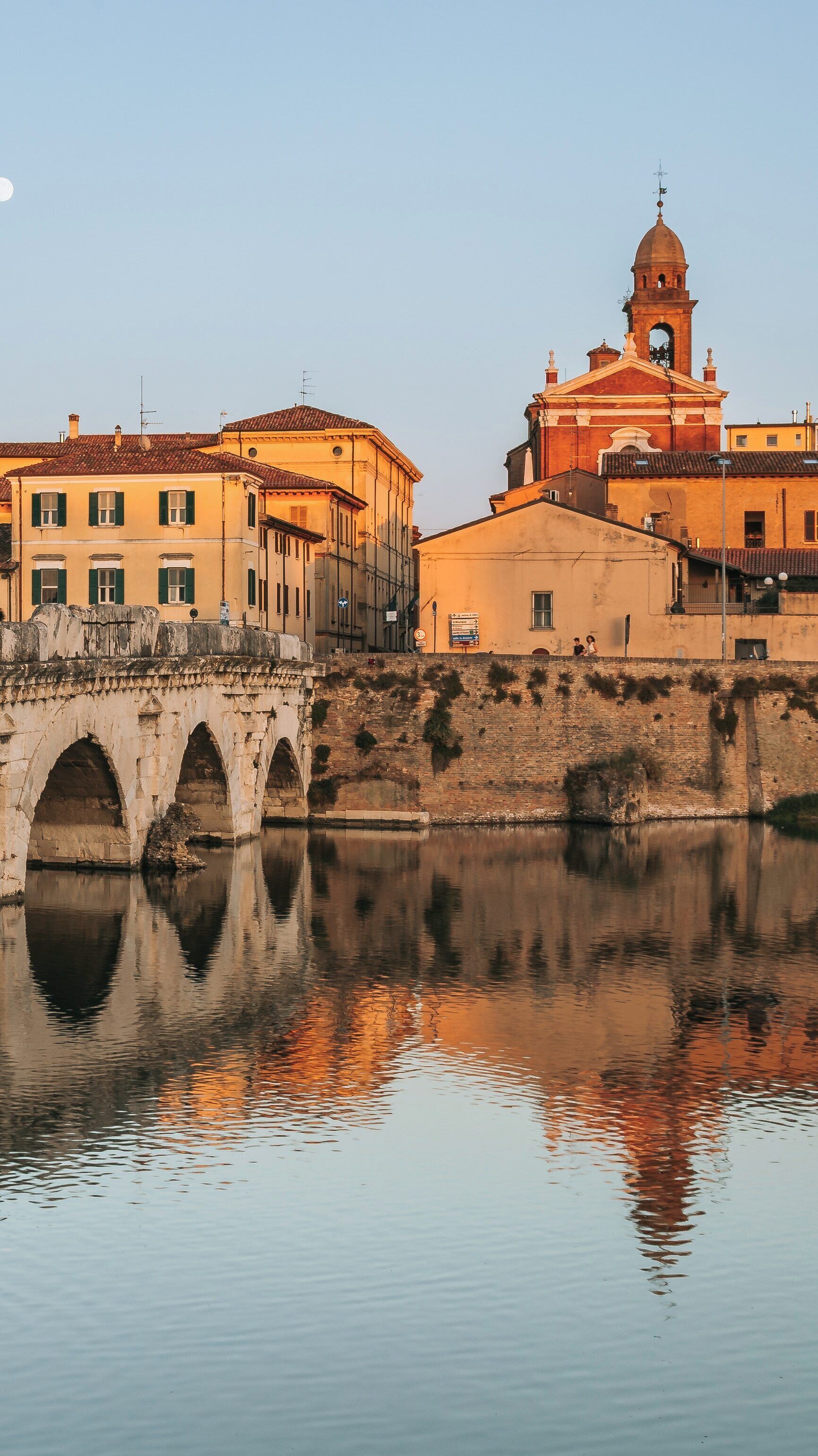 Historic Tiberius Bridge reflects in calm waters at sunset, overlooking charming buildings in San Giuliano a Mare, Rimini, Italy
