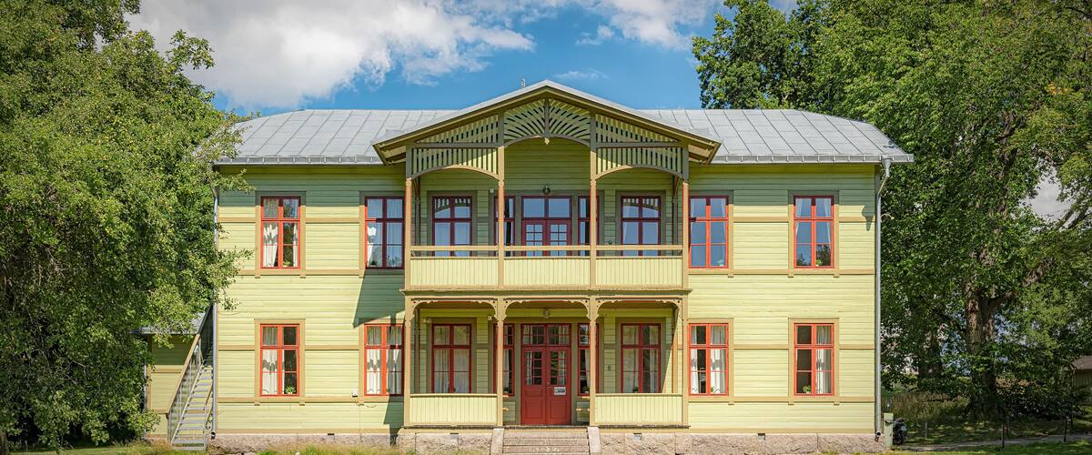 Un grand bâtiment à deux étages, doté d’une façade verte et de fenêtres à encadrement rouge, entouré d’une végétation luxuriante.