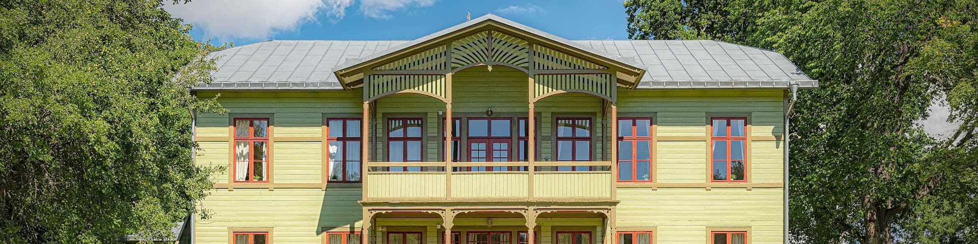 A large, two-story building with a green facade and red-framed windows, surrounded by lush greenery.