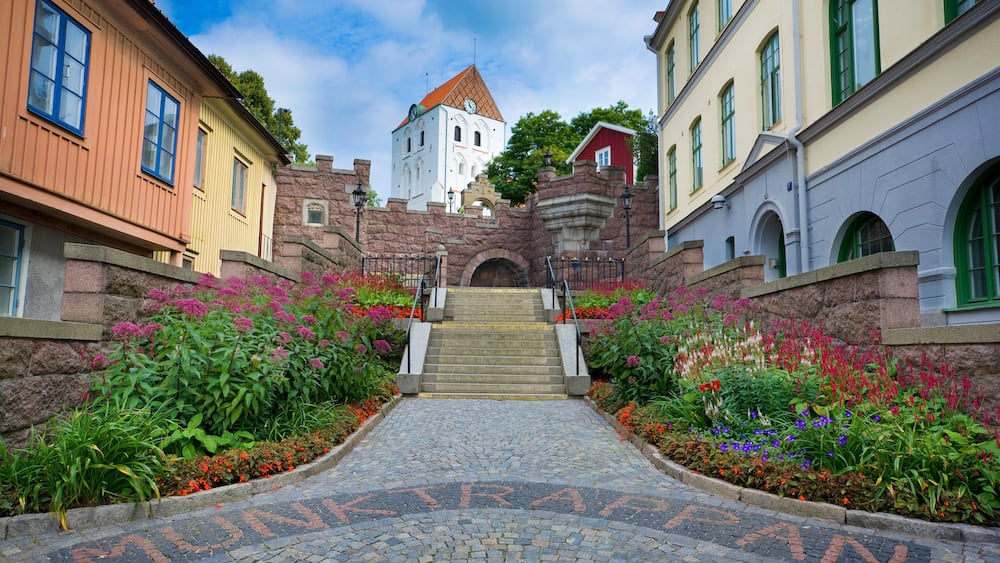 Ein Kopfsteinpflasterweg führt zu einem Turm mit einer Uhr, umgeben von bunten Blumen und historischen Gebäuden.
