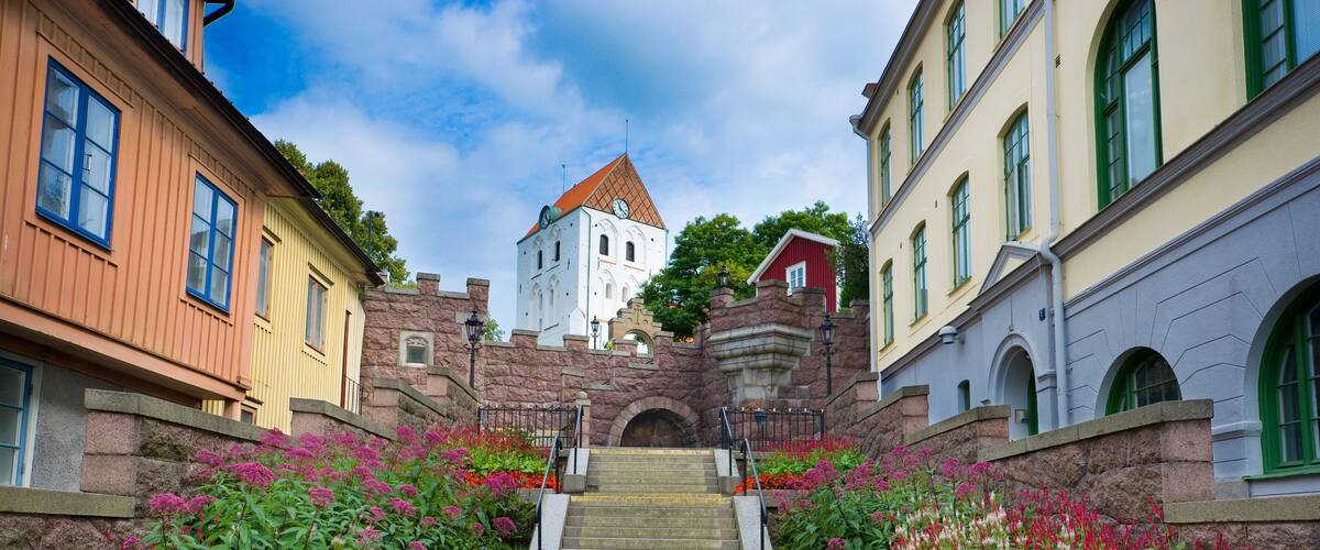 A cobblestone path leading to a tower with a clock, surrounded by colorful flowers and historic buildings.
