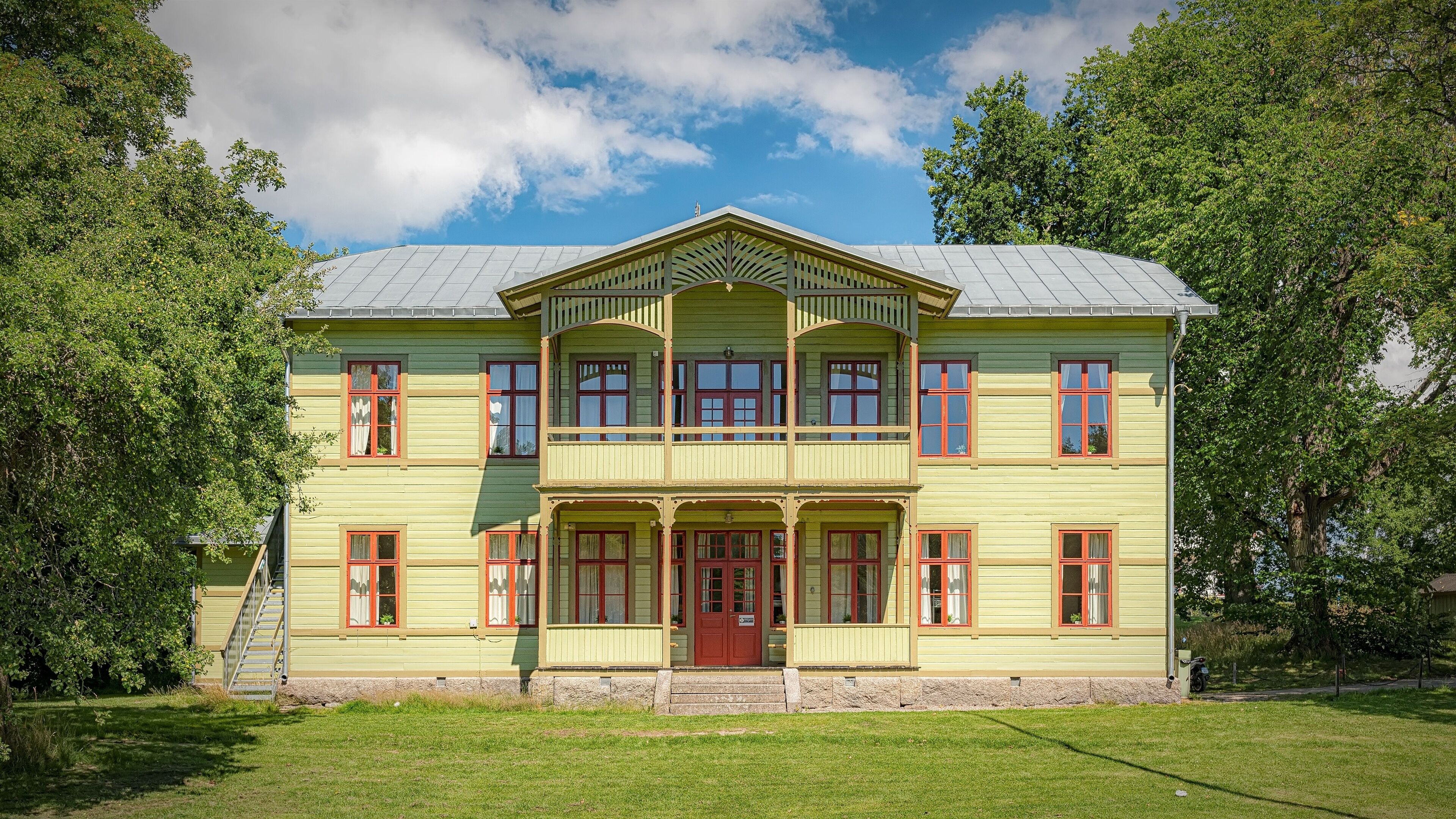 A large, two-story building with a green facade and red windows, surrounded by lush trees and a clear sky.