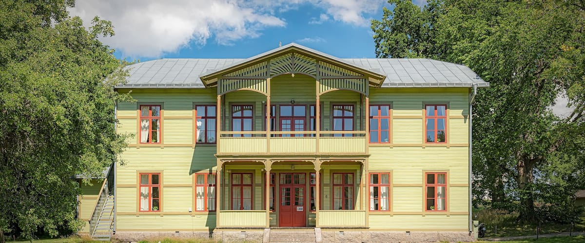 A large, two-story building with a green facade and red windows, surrounded by lush trees and a clear sky.