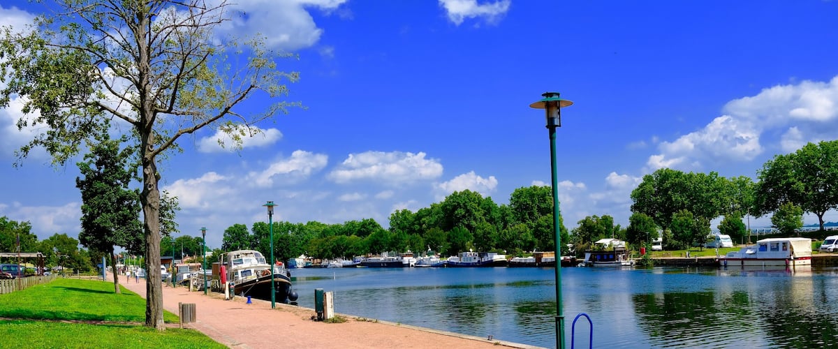 A riverside walkway with boats, trees, and a clear blue sky.