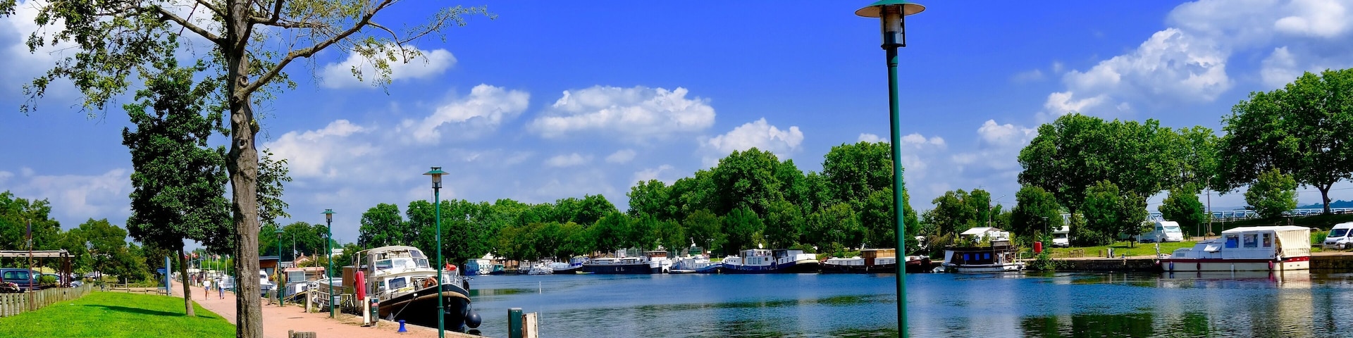 A riverside walkway with boats, trees, and a clear blue sky.