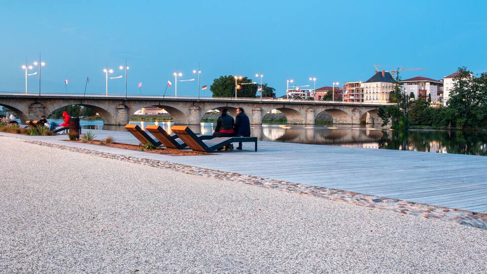 A riverside walkway with benches, a bridge, and buildings in the background.
