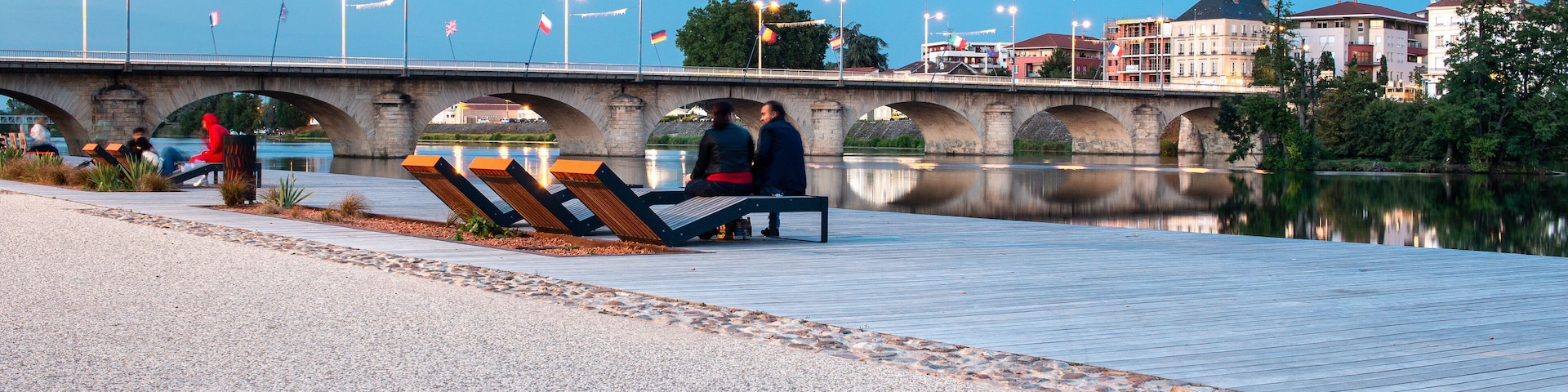 Uma passarela à beira do rio, com bancos, uma ponte e edifícios ao fundo.