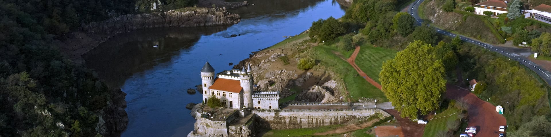 An aerial view of a castle on a river island, surrounded by lush greenery and a winding road.