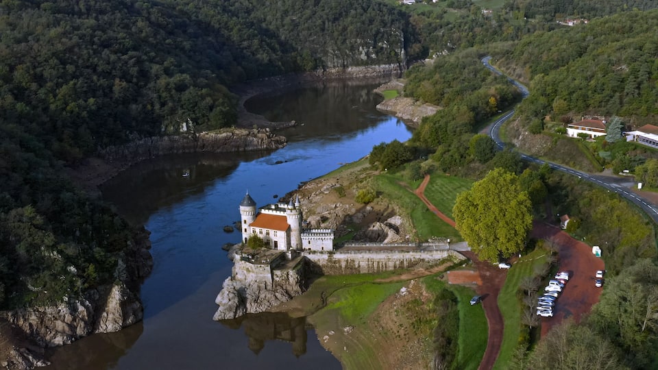An aerial view of a castle on a river island, surrounded by lush greenery and a winding road.