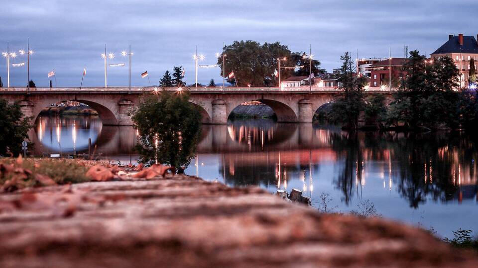 A bridge over a calm river with reflections of street lights and flags.