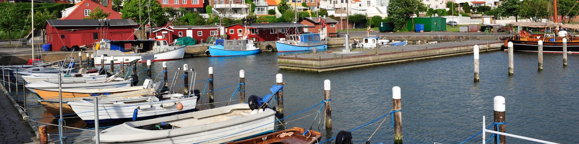 A harbor with various boats docked, a church with a tall steeple, and colorful houses in the background.