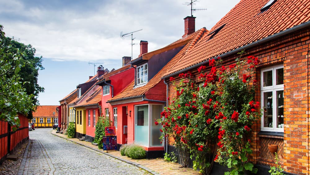 Une rue pavée bordée de maisons aux couleurs vives, de fleurs rouges et d’un ciel dégagé.