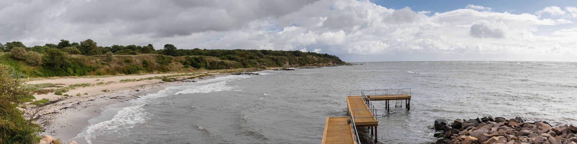 Un muelle de madera que se adentra en una costa rocosa, con olas rompiendo sobre la playa.