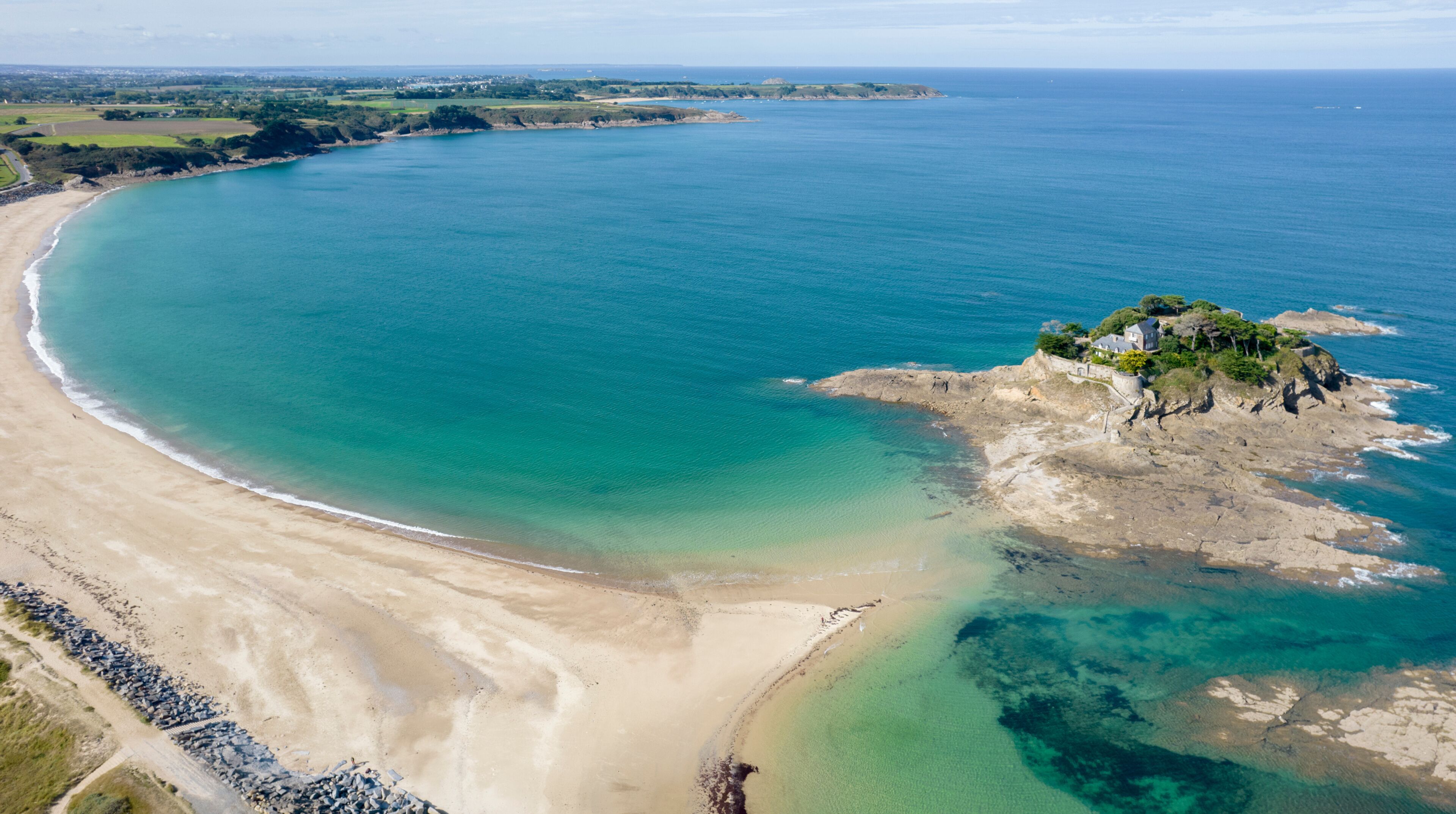Un paisaje costero con una playa arenosa, aguas turquesas cristalinas y un promontorio rocoso con vegetación.