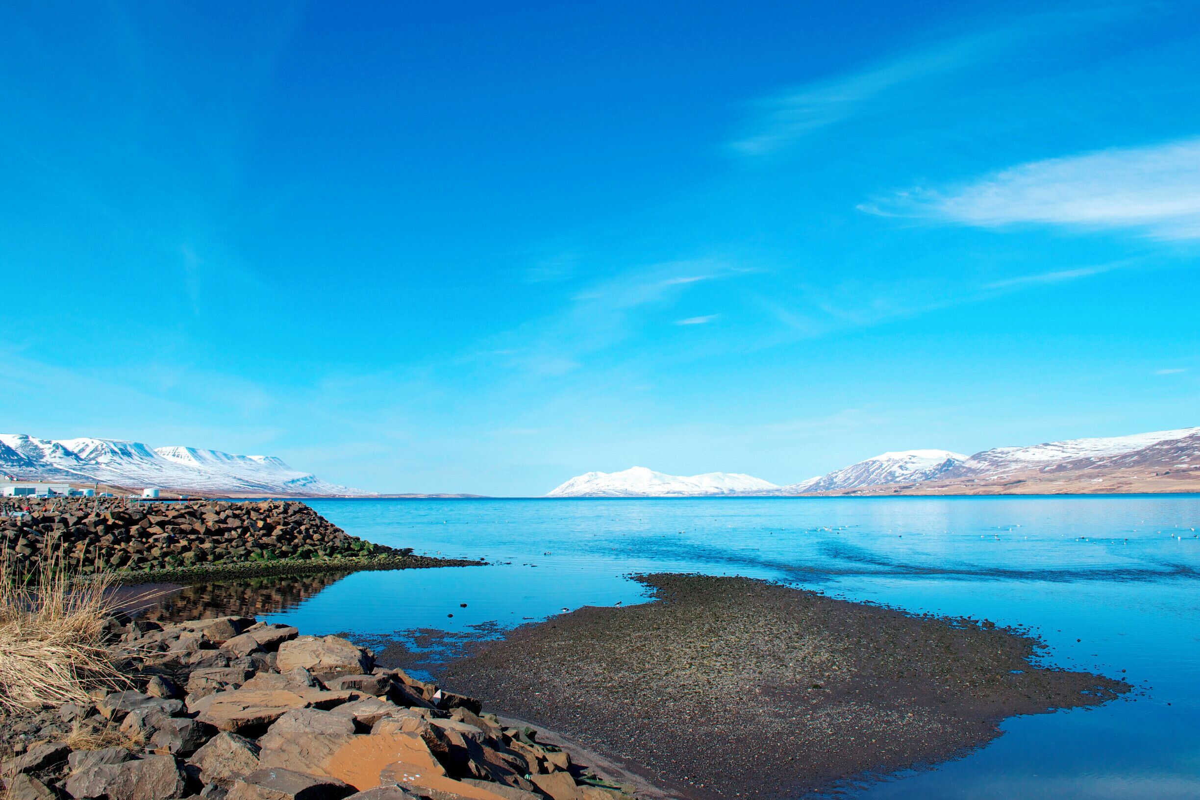 A calm lake with a rocky shoreline, clear blue sky, and snow-capped mountains in the background.