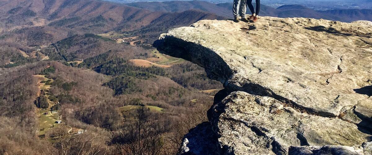 Two people standing on a rocky outcrop with a view of a valley and distant mountains.