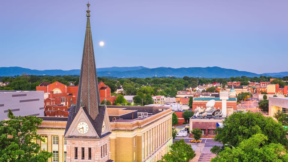 A town with a prominent church steeple, a clock tower, and a backdrop of mountains.