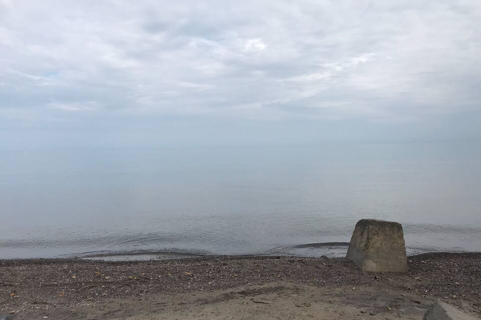 A pebbly beach with a concrete structure in the foreground and a calm sea under a cloudy sky.