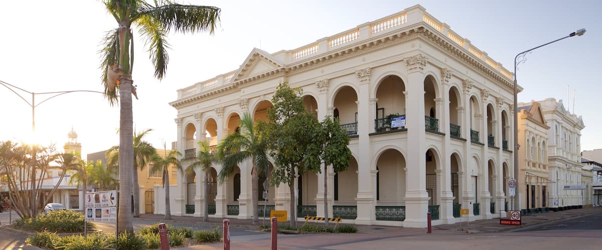 A historic building with arched windows and a balcony, surrounded by palm trees and a street.