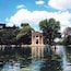 A tranquil lake with a classical pavilion, surrounded by lush trees and clear blue sky.