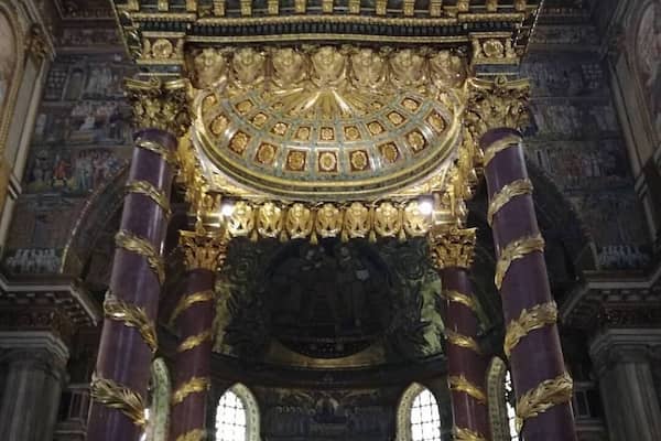 Un intérieur religieux richement décoré, avec des colonnes dorées et violettes, un plafond orné de motifs complexes et un autel central.