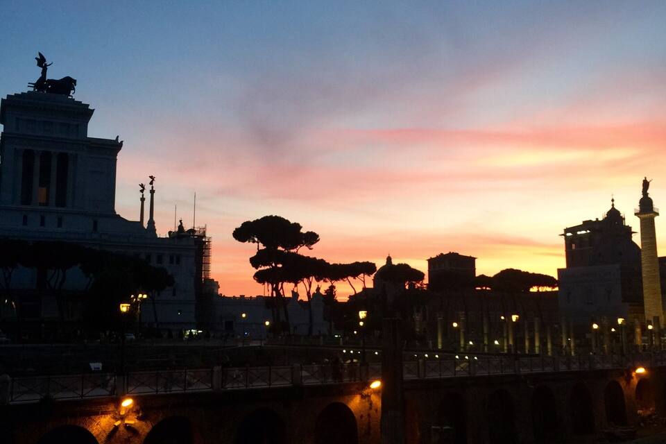 Um pôr do sol sobre uma ponte histórica com arcos, uma estátua e um edifício grande com colunas.