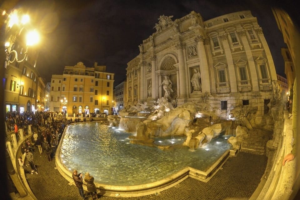 A nighttime view of the Trevi Fountain in Rome, Italy, with people around the water and the ornate Baroque building in the background.