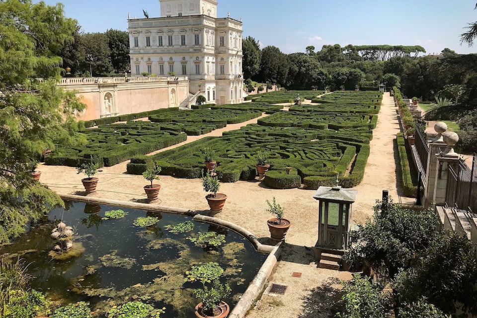 A formal garden with a central water feature, manicured hedges, and a large classical building in the background.