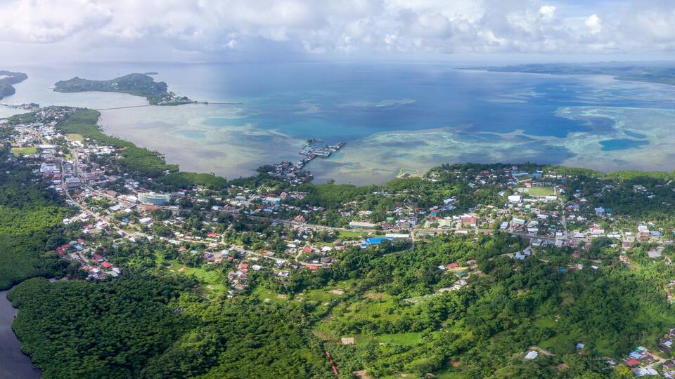 An aerial view of a coastal town with dense greenery, a pier extending into the sea, and multiple small islands in the background.