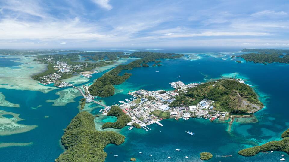 An aerial view of a tropical island with clear turquoise waters, dense green vegetation, and a small developed area with buildings and docks.