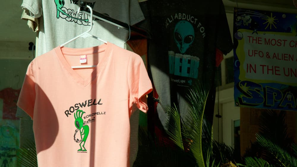 Ein pinkes T-Shirt mit einem grünen Alien-Aufdruck und Schrift, das in einem Laden zusammen mit anderer Ware ausgestellt ist.