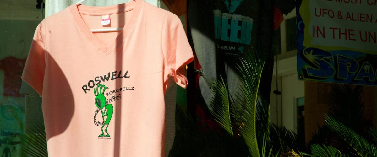 A pink t-shirt with a green alien graphic and text, hanging in a store with other merchandise.