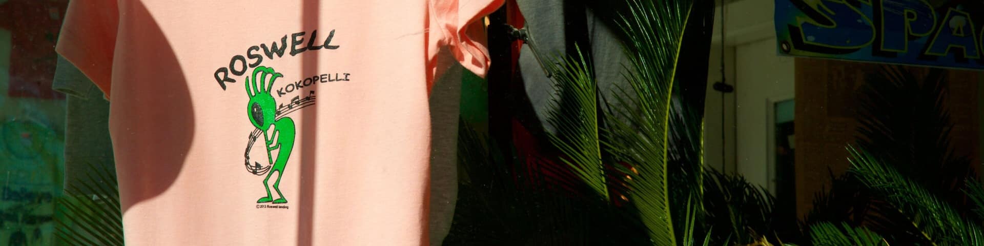 A pink t-shirt with a green alien graphic and text, hanging in a store with other merchandise.