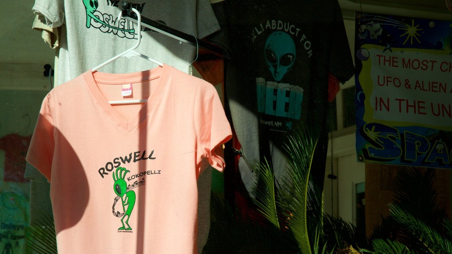 A pink t-shirt with a green alien graphic and text, hanging in a store with other merchandise.
