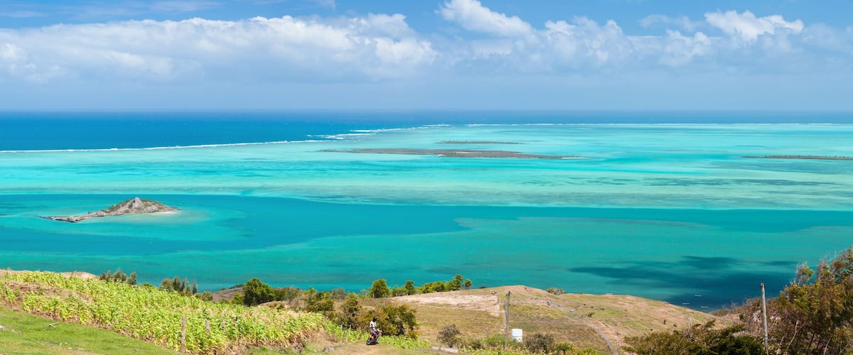 A coastal landscape with clear turquoise waters, a small island, and a hilly foreground with vegetation.