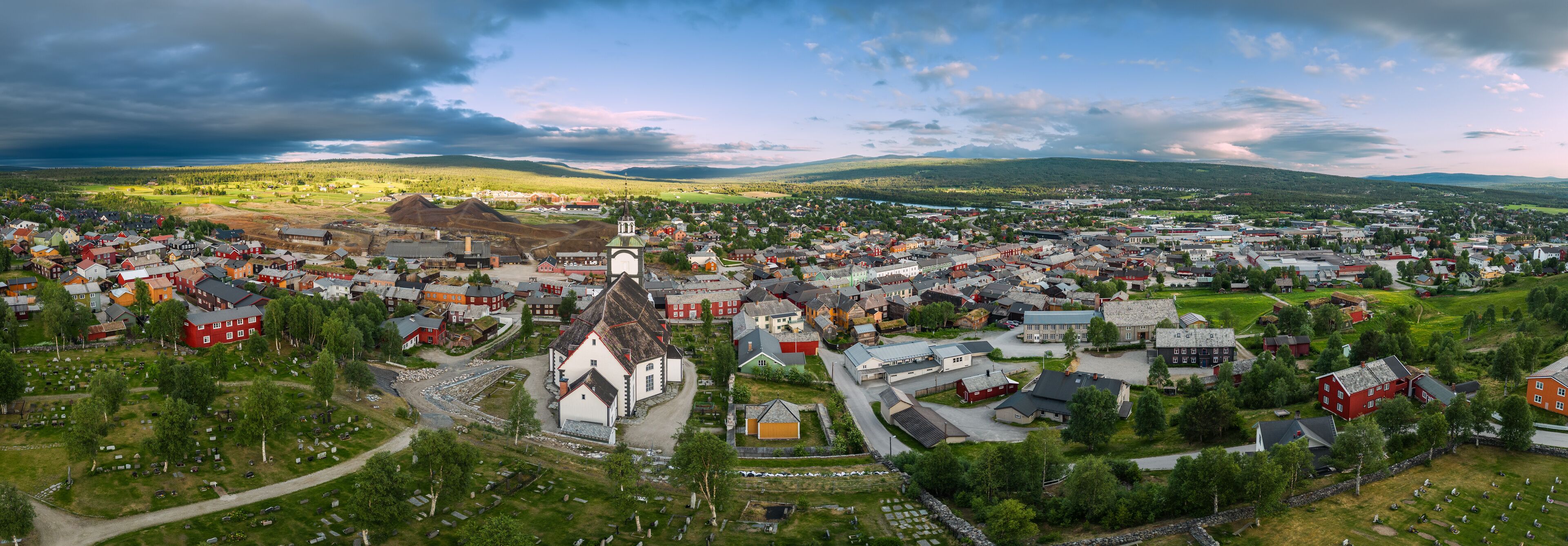 A sweeping drone panorama captures Roros, Norway, in summer splendor, with historic buildings nestled amid vibrant greenery under a dynamic sky