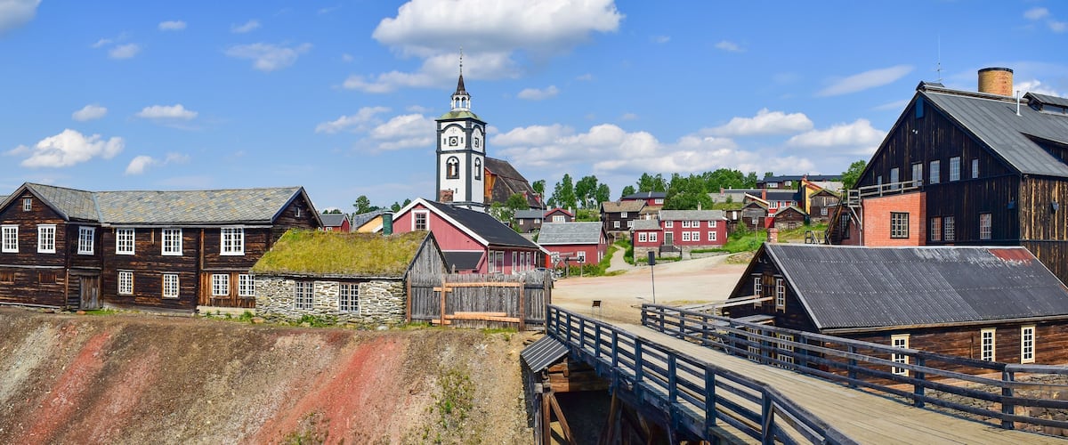 A historic town with a prominent clock tower, wooden buildings, and a wooden bridge.
