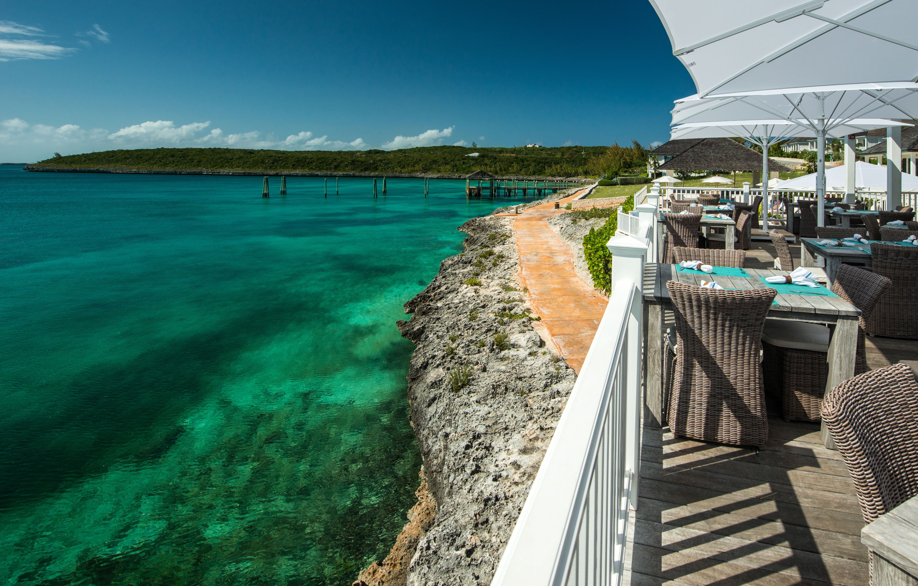 A coastal area with clear turquoise waters, a rocky shoreline, a pier extending into the sea, and a restaurant with outdoor seating overlooking the water.