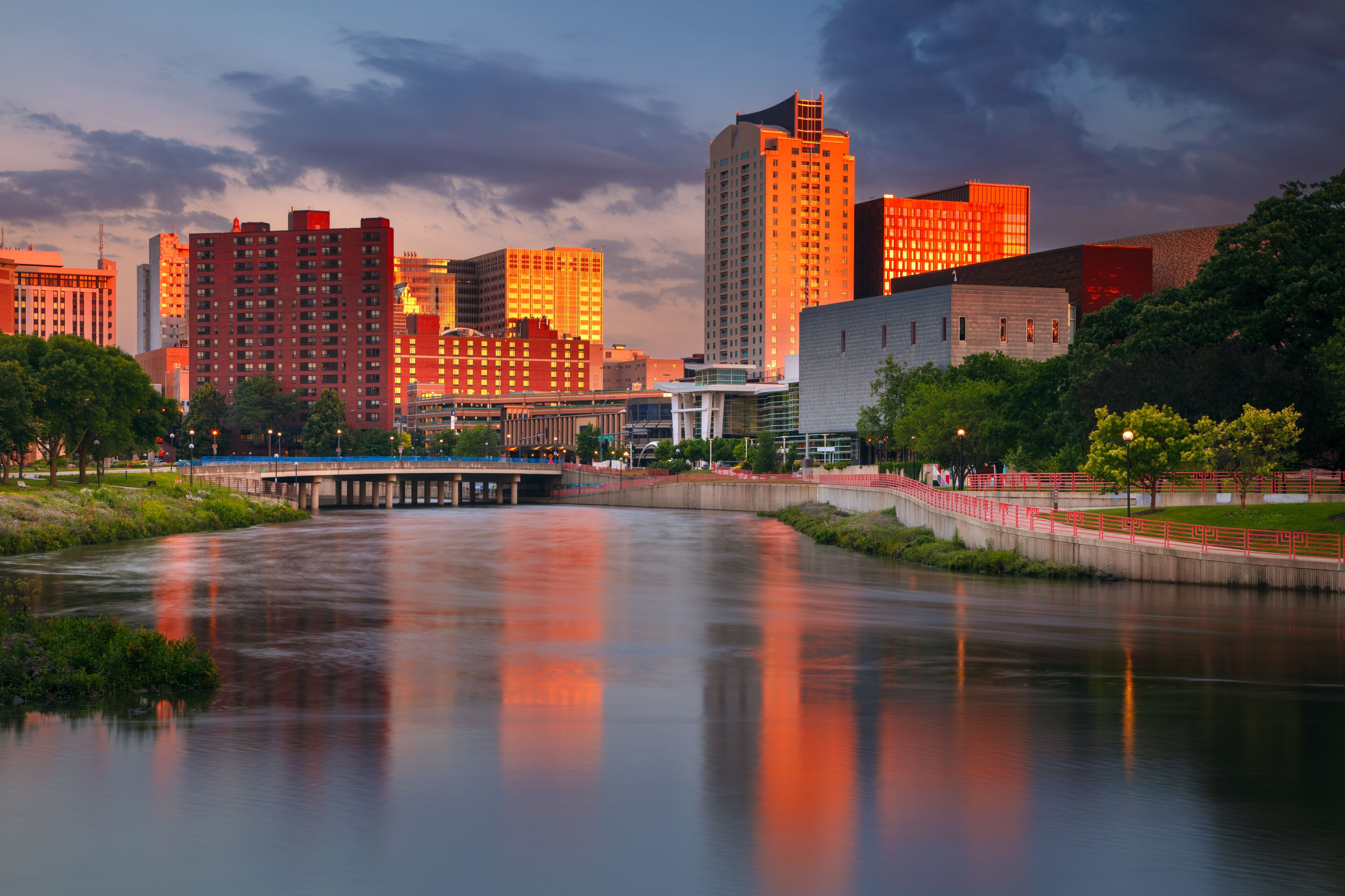 A river reflecting city buildings, a bridge, and a park with trees.