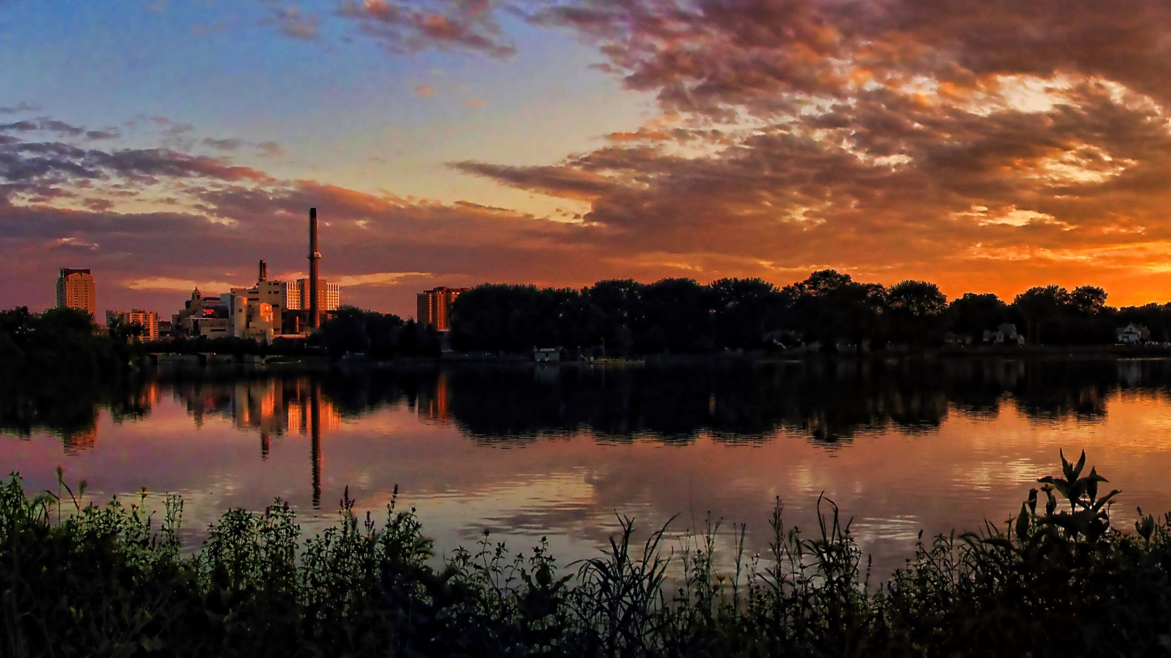 A sunset over a city skyline reflected in a calm lake, with industrial buildings and trees in the foreground.