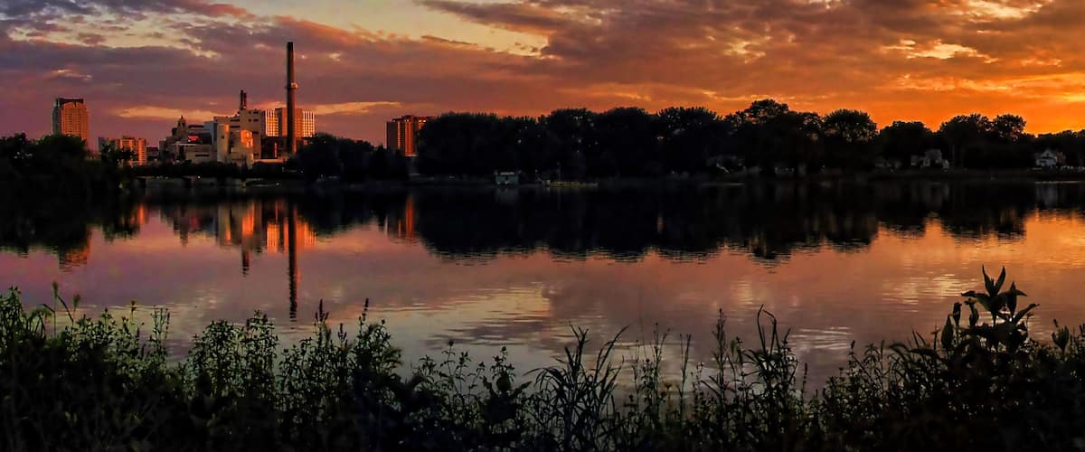 A sunset over a city skyline reflected in a calm lake, with industrial buildings and trees in the foreground.