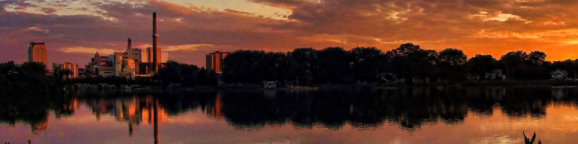 A sunset over a city skyline reflected in a calm lake, with industrial buildings and trees in the foreground.