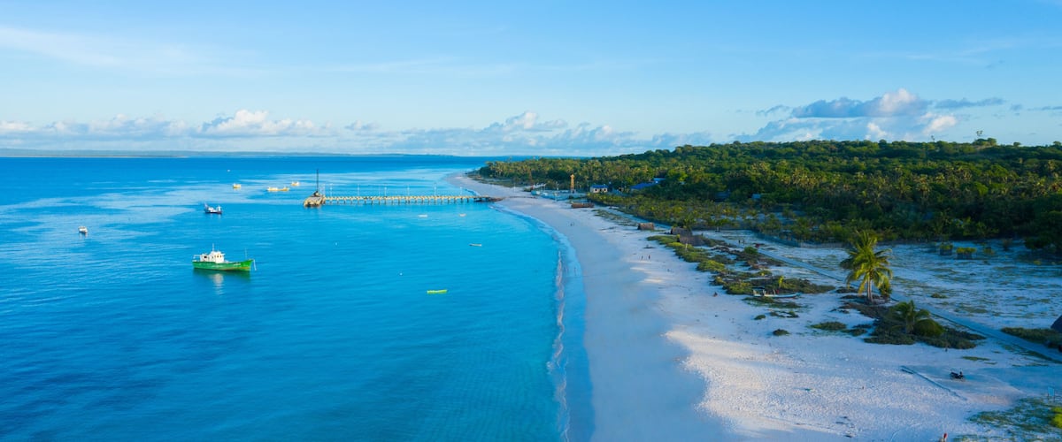 A coastal landscape with a sandy beach, clear blue waters, and a forested area.